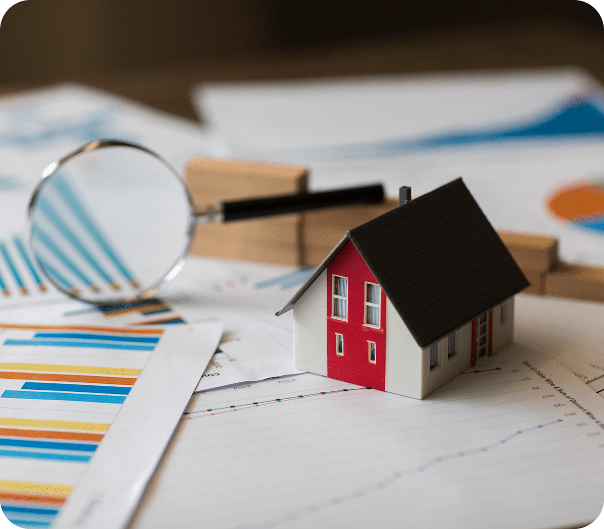 Miniature house on financial charts with magnifying glass.